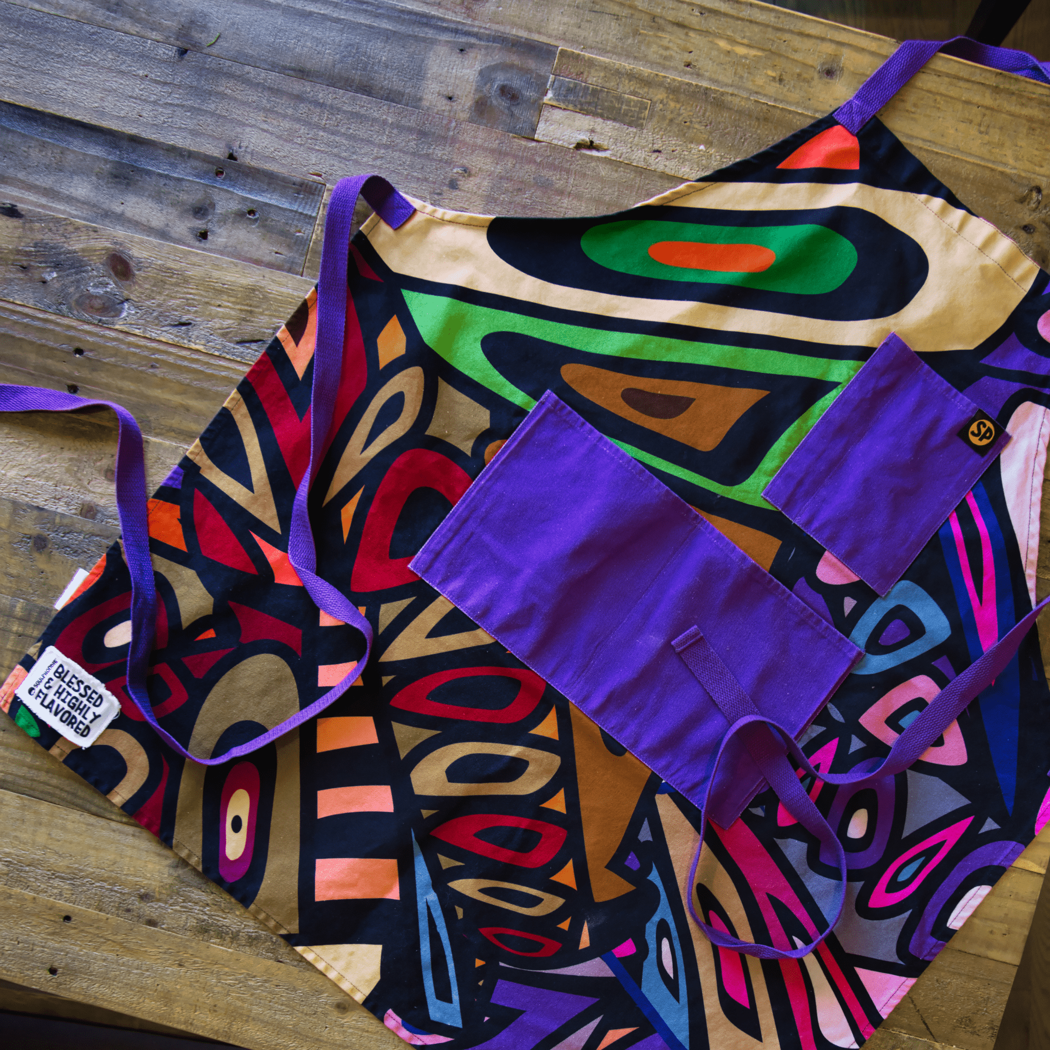Colorful abstract apron with purple straps and pockets on a wooden table, from Black-owned brand soulPhoodie.