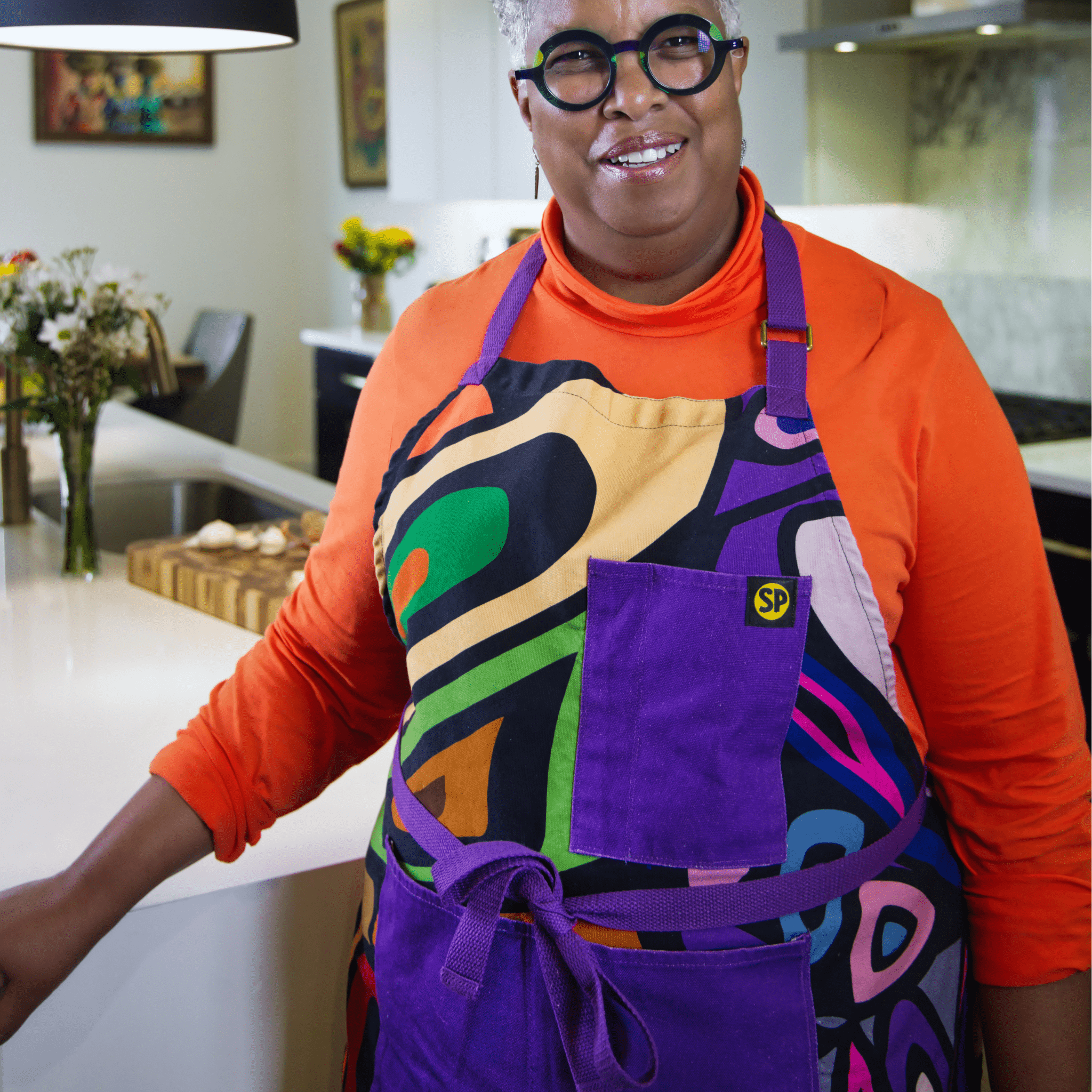 Woman wearing a multicolor abstract apron with purple straps from Black-owned brand soulPhoodie in a modern kitchen