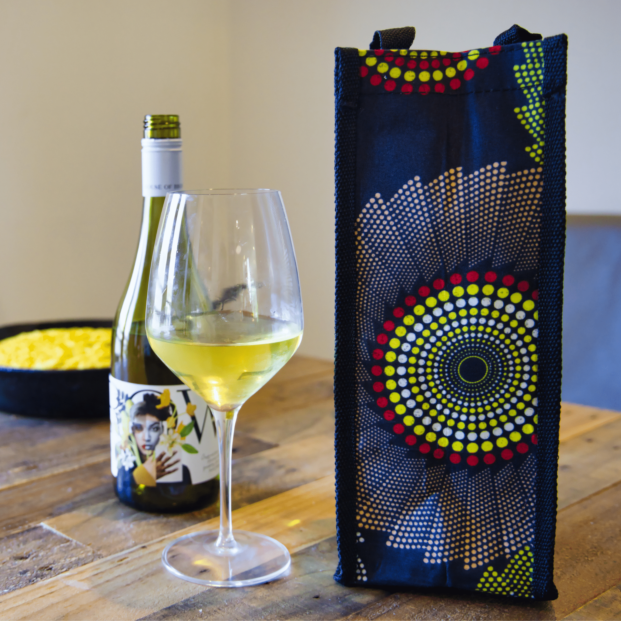 soulPhoodie wine bag with bold yellow and red circular patterns beside a glass of white wine and bottle on a wooden table.