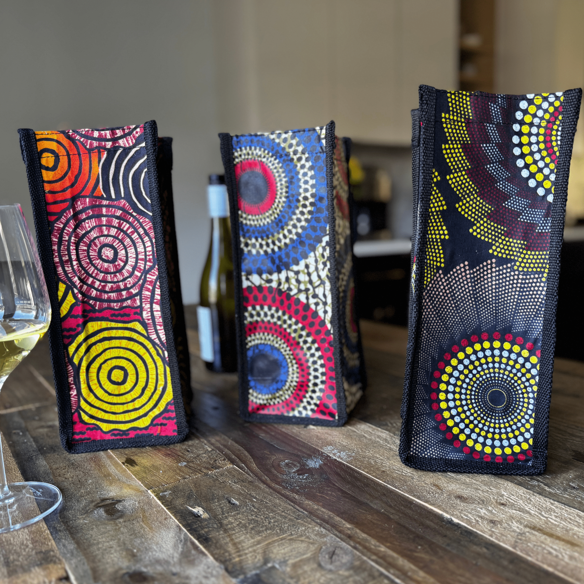 Three African-inspired wine bags from soulPhoodie featuring bold circular patterns and rich colors on a rustic wooden table