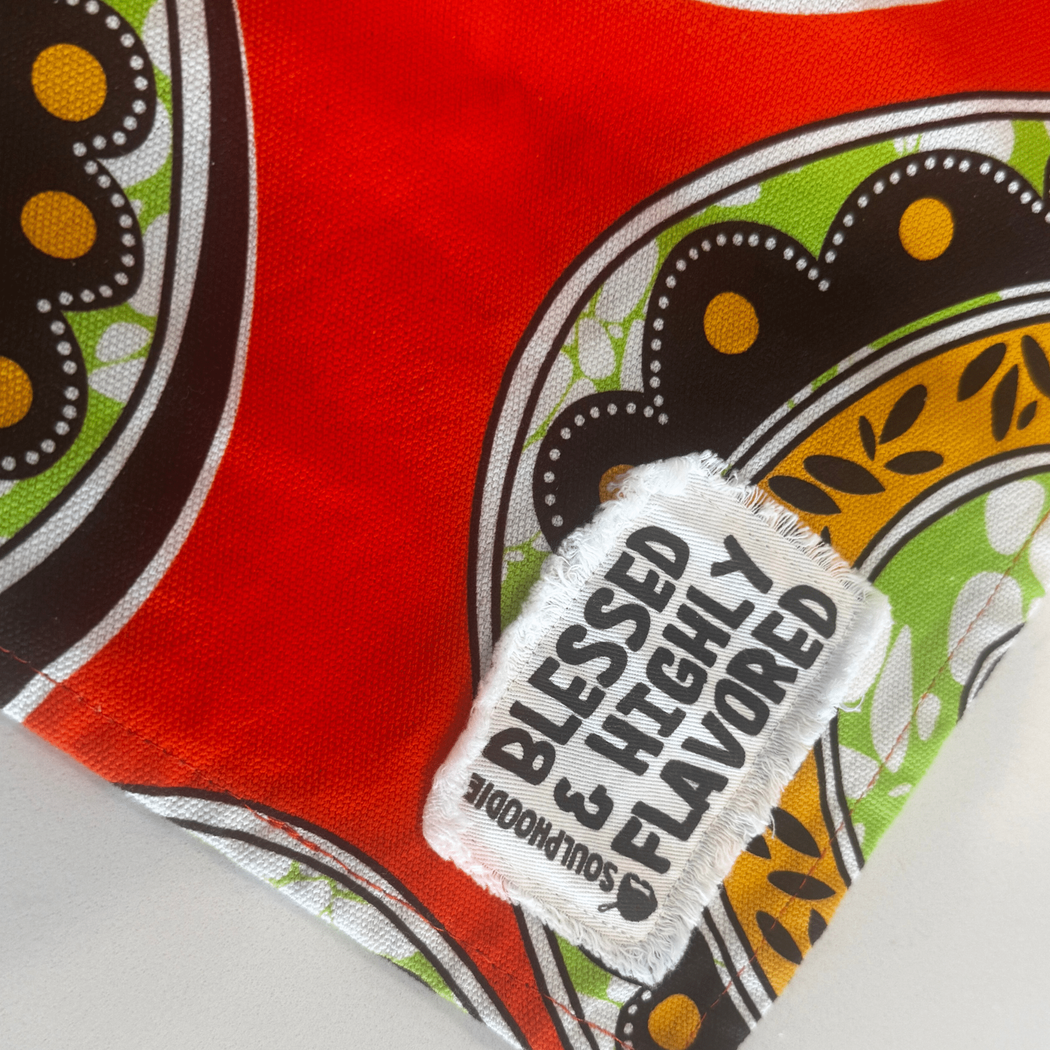 Close-up of the Blessed & Highly Flavored tag on the Susan Apron from soulPhoodie with bold orange and green fabric.